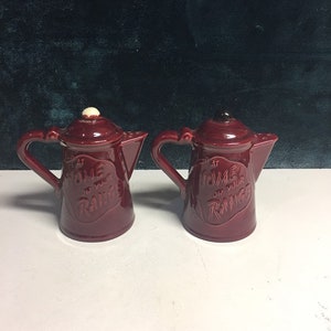 May include: Two red ceramic salt and pepper shakers shaped like coffee pots. The shakers have white and black tops and the text "At Home On Your Range" is printed on the front of each.