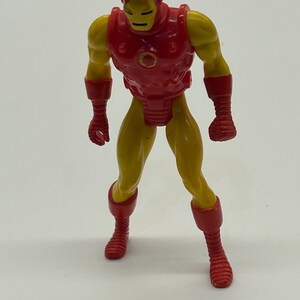 May include: A red and yellow plastic action figure of Iron Man. The figure is standing with its arms at its sides. The figure has a red chest plate with a circular opening in the center. The figure has a yellow body and red boots.