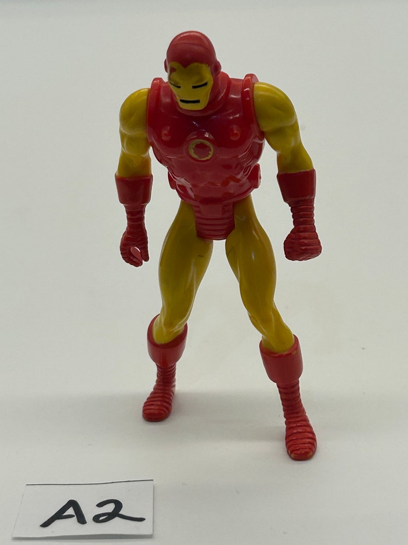 May include: A red and yellow plastic action figure of Iron Man. The figure is standing with its arms at its sides. The figure has a red chest plate with a circular opening in the center. The figure has a yellow body and red boots.
