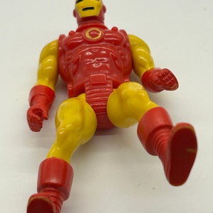 May include: A vintage action figure of Iron Man in a red and yellow suit. The figure is posed with one leg raised and the other leg extended. The figure has a detailed head sculpt with a red and yellow helmet.