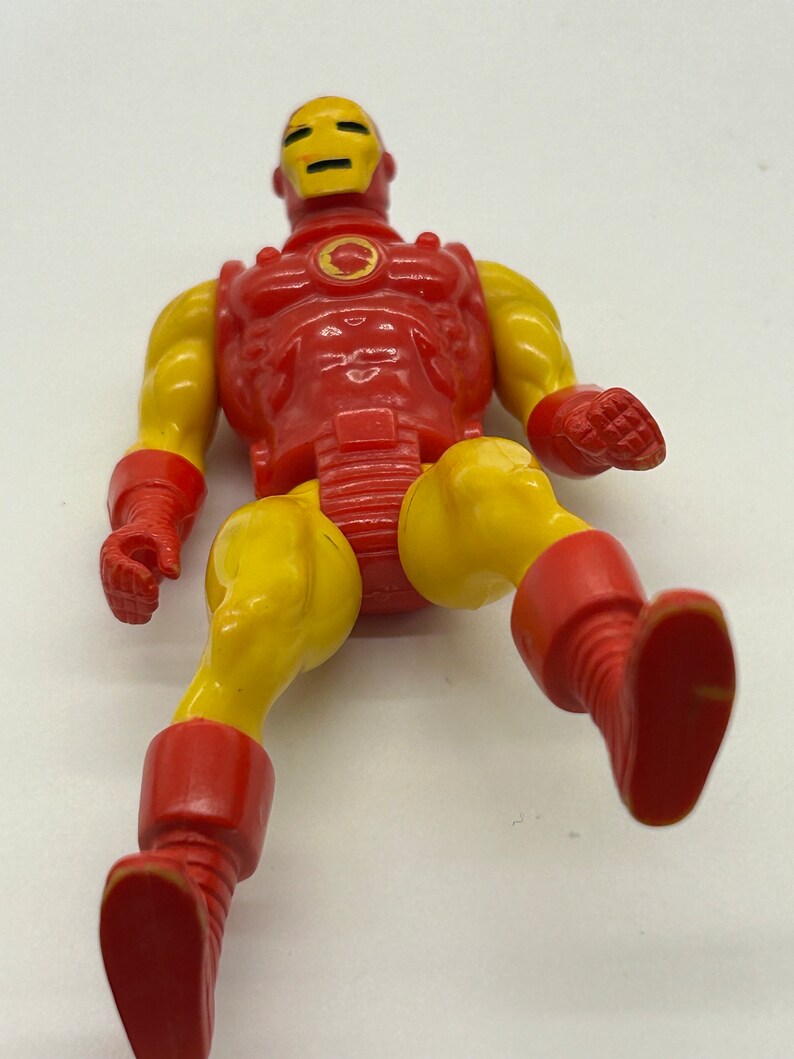 May include: A vintage action figure of Iron Man in a red and yellow suit. The figure is posed with one leg raised and the other leg extended. The figure has a detailed head sculpt with a red and yellow helmet.