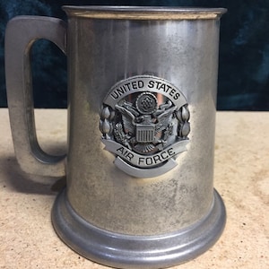 May include: A silver metal beer stein with a United States Air Force emblem. The emblem features a bald eagle with a shield and the words "United States Air Force".