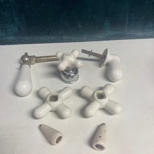 May include: A set of eight vintage white porcelain faucet handles, including two with cross-shaped knobs, two with round knobs, and four with long, pointed handles. One of the cross-shaped knobs has the word "HOT" printed on it.