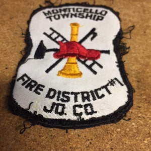 May include: White embroidered patch with black trim. It features a red fire hat with a yellow trumpet, crossed ladders, and an axe. The text reads "Monticello Township Fire District #1 Jo Co."