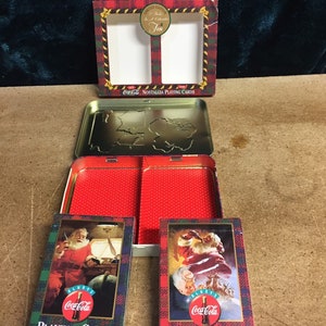 May include: Two decks of Coca-Cola playing cards in a collectible tin. The tin is red and has a Coca-Cola logo on it. The cards feature a Santa Claus illustration with the text "Always Coca-Cola" and "Playing Cards".