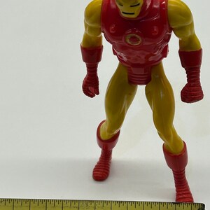 May include: A red and yellow plastic action figure of Iron Man. The figure is standing on a white surface with a ruler in the foreground. The ruler is marked with inches and has the text "1/16 ft" and "1976" printed on it.