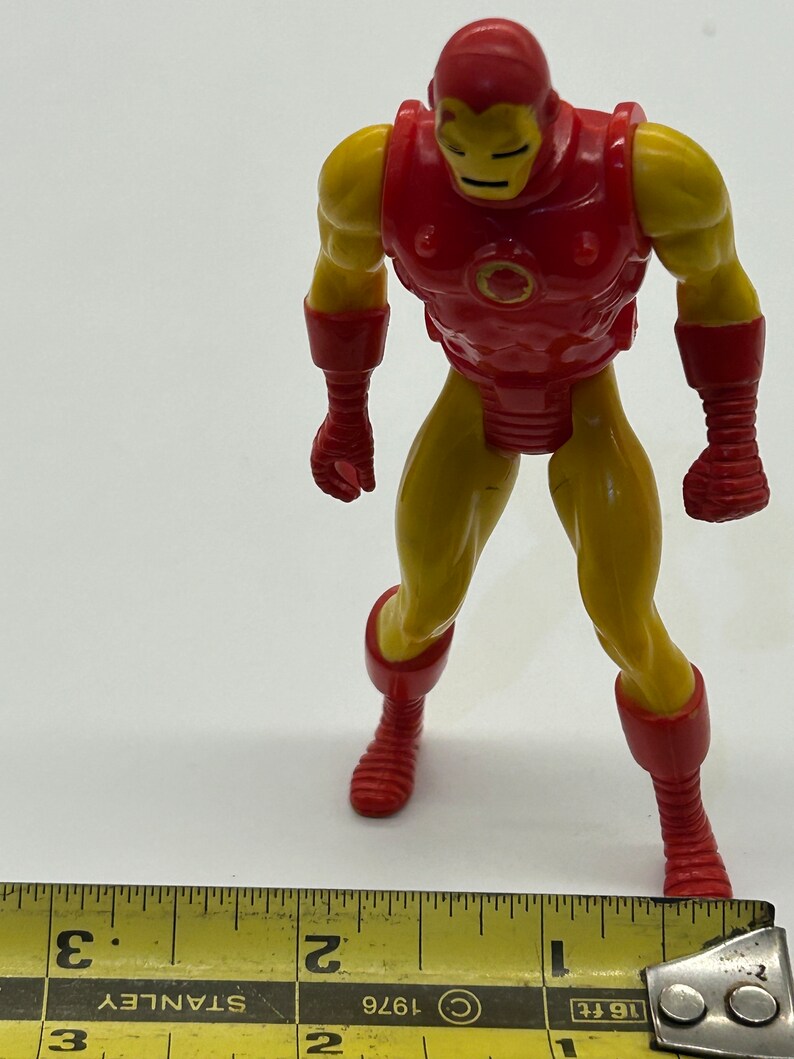 May include: A red and yellow plastic action figure of Iron Man. The figure is standing on a white surface with a ruler in the foreground. The ruler is marked with inches and has the text "1/16 ft" and "1976" printed on it.