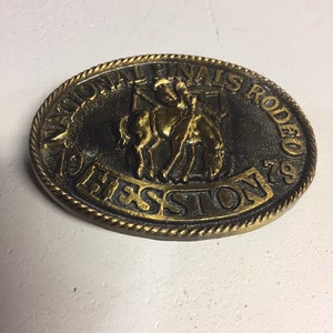 May include: A gold-toned belt buckle with a rope-like border. The buckle features a depiction of a cowboy riding a horse, with the text "National Finals Rodeo" and "1979" around the edge.
