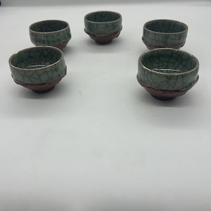 May include: Six small green teacups with a crackle glaze and gold accents. The cups are arranged in a circle on a white surface.