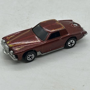 May include: A vintage die-cast toy car in a metallic burgundy finish. The car features black wheels, a black roof, and gold and yellow accent stripes on the hood and roof. The car has a detailed front grill and a circular design on the rear.