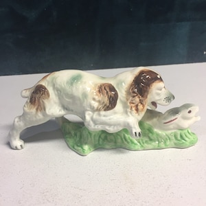 May include: A ceramic figurine of a brown and white dog chasing a fish. The dog is running on a green base.