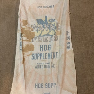 May include: A vintage white burlap sack with brown stains. The sack is labeled "100 LBS. NET" and features a logo for "WAYNE FEEDS" with a horse and rider. The text "HOG SUPPLEMENT" is printed in large letters, along with the manufacturer's name, "ALLIED MILLS INC.". The sack also has the text "HOG SUPP." printed on it.
