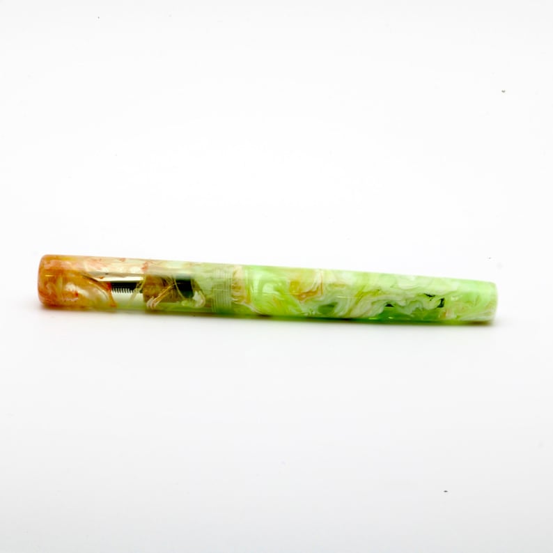 May include: A colorful fountain pen with a marbled design. The pen features a blend of orange, yellow, and green hues. The nib is visible, and the pen is set against a white background. A writing instrument.
