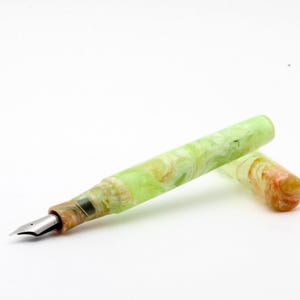 May include: A fountain pen with a marbled design in shades of green, yellow, and brown. The pen has a silver nib and a matching cap. The pen is lying on a white surface.