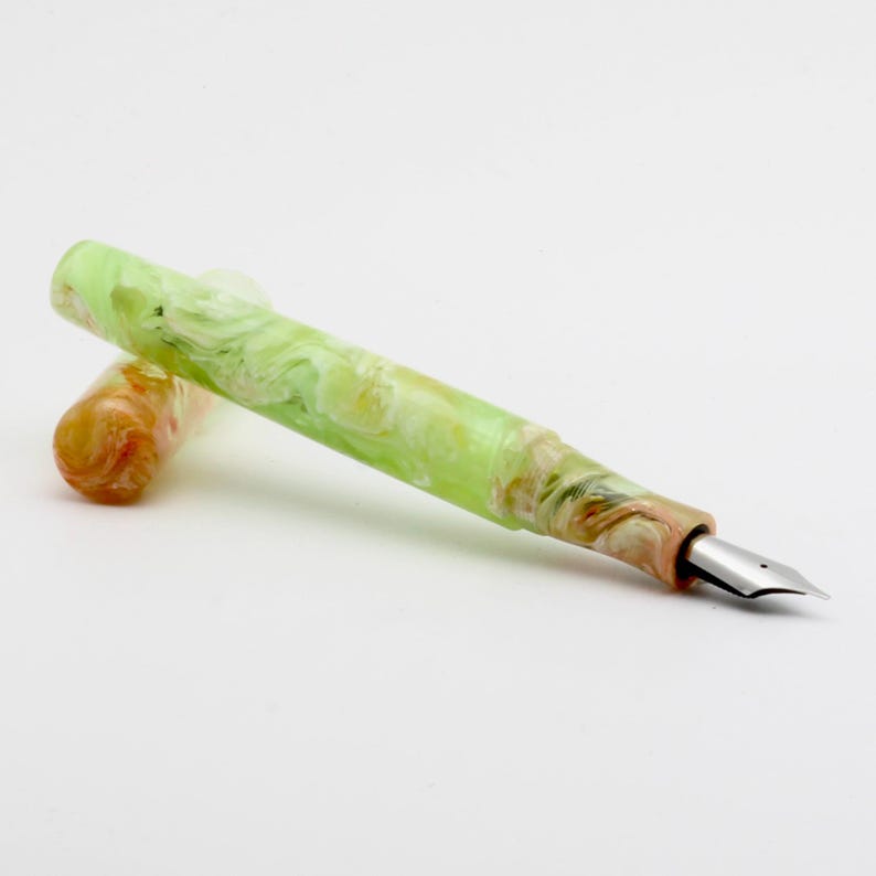 May include: A fountain pen with a marbled design in shades of green, yellow, and brown. The pen has a silver nib and a matching cap. The pen is shown on a white background.