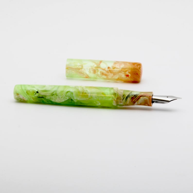 May include: A fountain pen with a marbled design in shades of green, white, and brown. The pen's cap is off, revealing the nib. The pen is made of a translucent material.