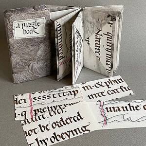 May include: A gray and white marbled book titled "a puzzle book" is open to reveal pages with black calligraphy.  The book is surrounded by several smaller pieces of paper with black calligraphy, including the words "not be ordered" and "Thy obeying".