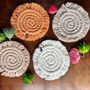 May include: Four handmade crocheted coasters in beige, white, and orange with fringe detailing. The coasters are arranged on a wooden surface.