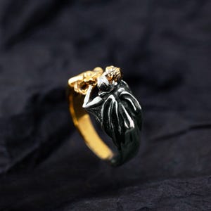 May include: A two-tone ring with a gold band and a dark silver design. The ring features a detailed figure with a gold head and a dark silver body. The ring is set against a dark background.