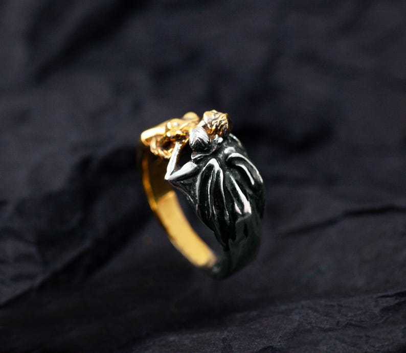 May include: A two-tone ring with a gold band and a dark silver design. The ring features a detailed figure with a gold head and a dark silver body. The ring is set against a dark background.
