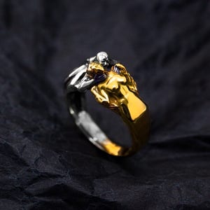 May include: A two-tone ring featuring a gold-colored figure embracing a silver-colored figure. The ring has a detailed design with a dark background.