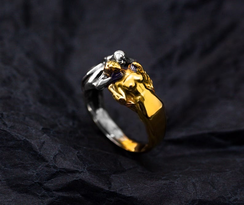 May include: A two-tone ring featuring a gold-colored figure embracing a silver-colored figure. The ring has a detailed design with a dark background.