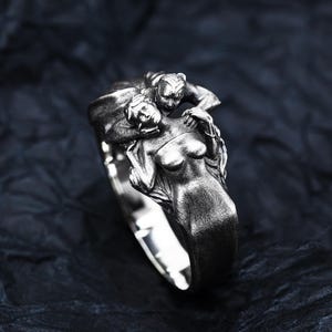 May include: A silver ring featuring a sculpted design of two figures intertwined. One figure is a woman, and the other appears to be a creature. The ring has a polished finish and is set against a dark background.