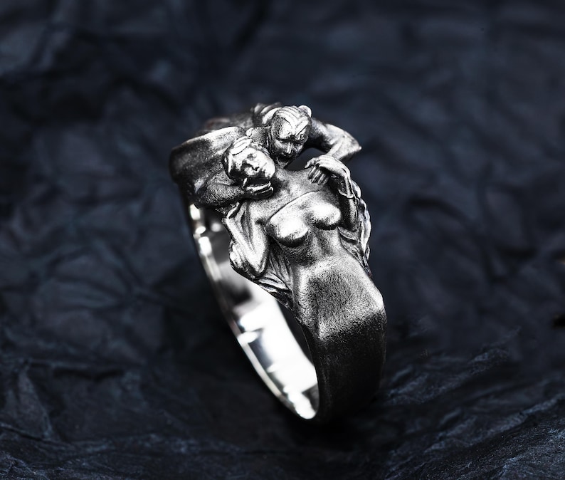 May include: A silver ring featuring a sculpted design of two figures intertwined. One figure is a woman, and the other appears to be a creature. The ring has a polished finish and is set against a dark background.