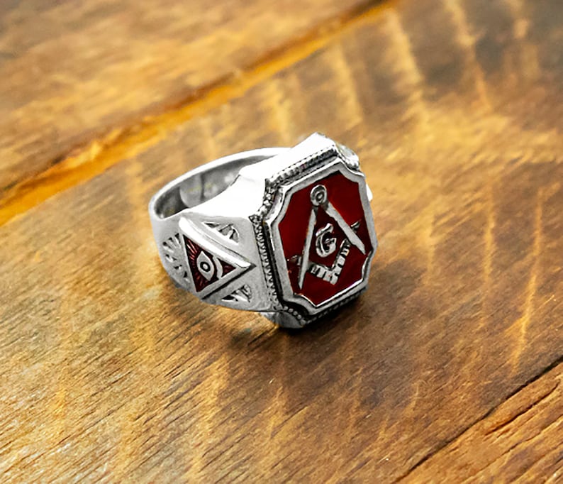 Op de afbeelding: Een zilveren vrijmetselaarsring met een rode emaille-inleg met een passer en een winkelhaak, en een kleiner alziend oogontwerp aan de zijkant.