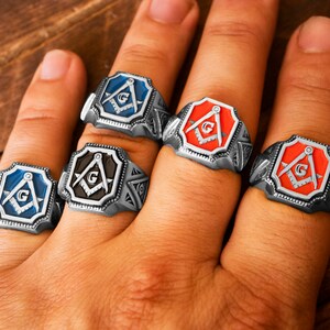 Op de afbeelding: Vier zilveren ringen met elk een vrijmetselaars symbool. De ringen hebben een zwart, blauw, rood en oranje emaille ontwerp.