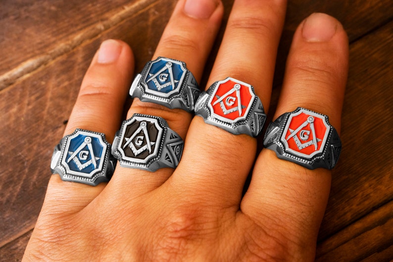Op de afbeelding: Vier zilveren ringen met elk een vrijmetselaars symbool. De ringen hebben een zwart, blauw, rood en oranje emaille ontwerp.