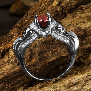 Op de afbeelding: Een zwarte ring met een rode hartvormige edelsteen en schedeldetails. De ring is bezet met kleine witte edelstenen.