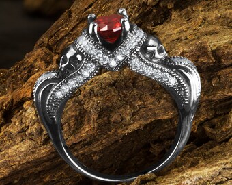 Puede incluir: Un anillo negro con una piedra preciosa roja en forma de corazón y detalles de calavera. El anillo está engastado con pequeñas piedras blancas.