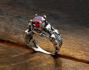 Puede incluir: Un anillo de plata con una piedra preciosa roja engastada en un diseño de calavera.