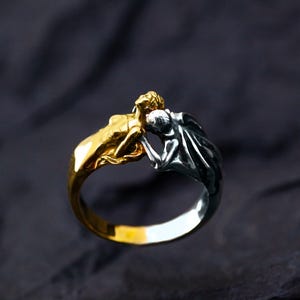 May include: A two-tone ring featuring two figures in a close embrace. One figure is gold-toned, the other silver-toned. The ring has a polished finish and is set against a dark background. The ring is a unique piece of jewelry.