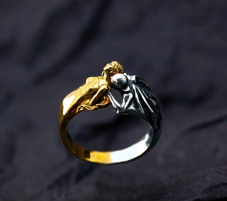 May include: A two-tone ring featuring two figures in a close embrace. One figure is gold-toned, the other silver-toned. The ring has a polished finish and is set against a dark background. The ring is a unique piece of jewelry.