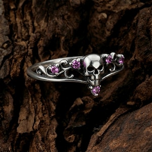 Könnte beinhalten: Ein silberfarbener Ring mit einem zentralen Totenkopfmotiv, das von geschwungenen Metallverzierungen flankiert wird. Das Design ist mit fünf kleinen, runden, leuchtend pinkfarbenen Edelsteinen besetzt, die um den Totenkopf und an den Seiten sitzen.