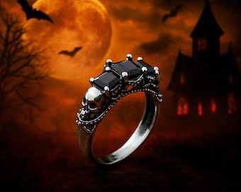 Puede incluir: Un anillo plateado con tres grandes piedras negras cuadradas. El anillo presenta un diseño de calavera en el lateral y está ambientado en un fondo de Halloween con luna llena, murciélagos y una casa encantada.