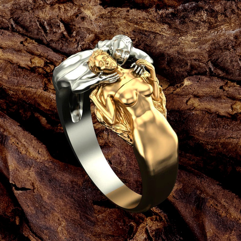 May include: A silver and gold ring featuring a couple in an embrace. The man is positioned above the woman, with his arms around her neck. The woman's hair flows behind her. The ring is displayed against a wooden backdrop.