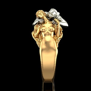 May include: A gold ring featuring a detailed sculpture of a woman being embraced by a man. The woman is wearing a flowing gown and the man is holding her close. The ring is a statement piece with a unique design.