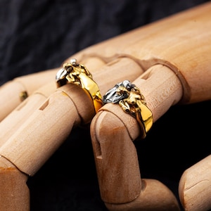 May include: Two gold-colored rings with a detailed lion head design are displayed on a wooden hand. One ring has a silver-colored mane. The rings are set against a dark background, highlighting the jewelry's intricate details.