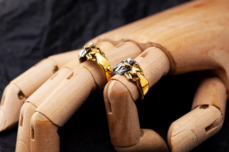 May include: Two gold-colored rings with a detailed lion head design are displayed on a wooden hand. One ring has a silver-colored mane. The rings are set against a dark background, highlighting the jewelry's intricate details.