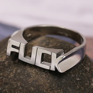 May include: A silver ring with the word "FUCK" spelled out in raised letters.