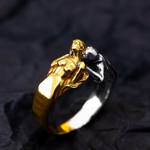 May include: A two-tone ring featuring a gold-colored figure embracing a silver-colored figure. The ring has a detailed design with a smooth, polished finish. The figures are sculpted in a stylized, artistic manner, creating a unique piece of jewelry.