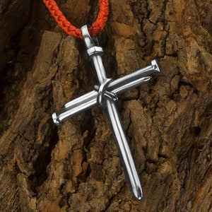 Puede incluir: Un colgante de cruz de plata con clavos, colgado de un cordón rojo. La cruz está hecha de metal y tiene un acabado martillado. El colgante es un símbolo religioso y es una pieza de joyería popular.
