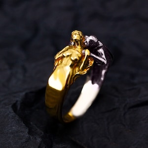 May include: A gold and silver ring featuring two figures embracing. One figure is gold, the other is silver. The ring has a detailed design, with the figures' forms clearly visible. The ring is set against a dark background.