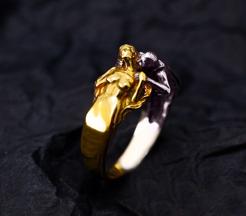 May include: A gold and silver ring featuring two figures embracing. One figure is gold, the other is silver. The ring has a detailed design, with the figures' forms clearly visible. The ring is set against a dark background.