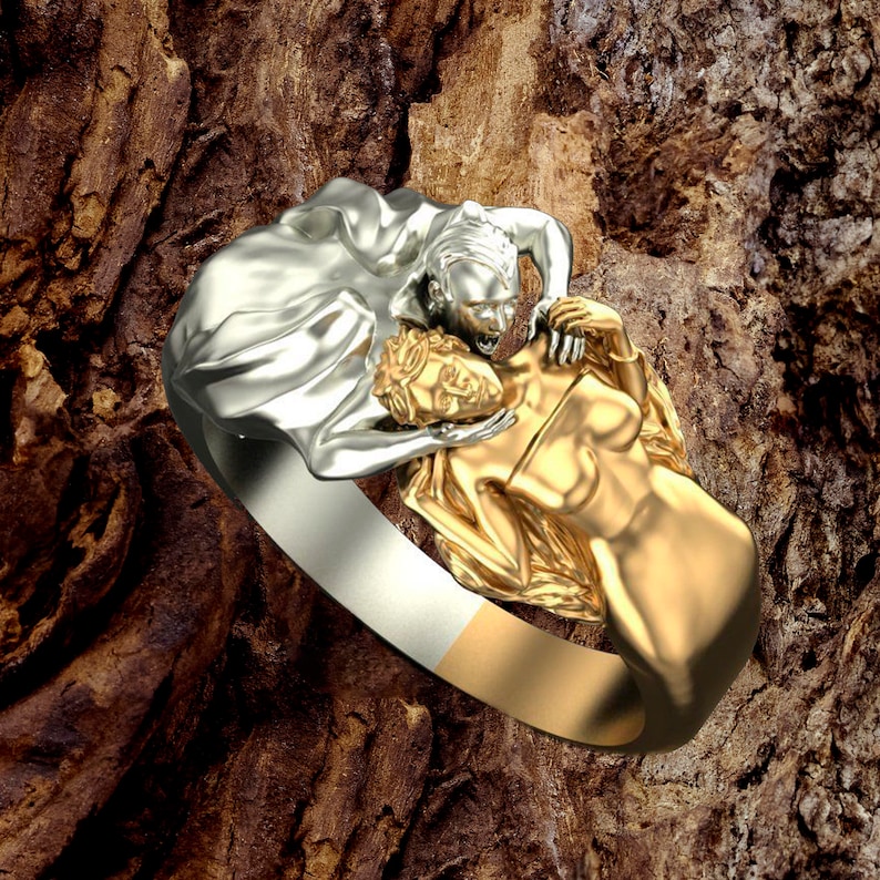 May include: A silver and gold ring featuring a sculpted design of two figures embracing. The silver figure is on top of the gold figure, and their faces are visible.