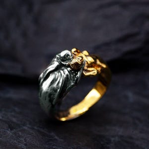 May include: A two-tone ring with a detailed sculpture. The ring features a silver-colored figure on top of a gold-colored band. The sculpture appears to be a person or creature with intricate details. The ring is set against a dark background.
