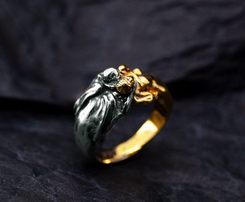 May include: A two-tone ring with a detailed sculpture. The ring features a silver-colored figure on top of a gold-colored band. The sculpture appears to be a person or creature with intricate details. The ring is set against a dark background.